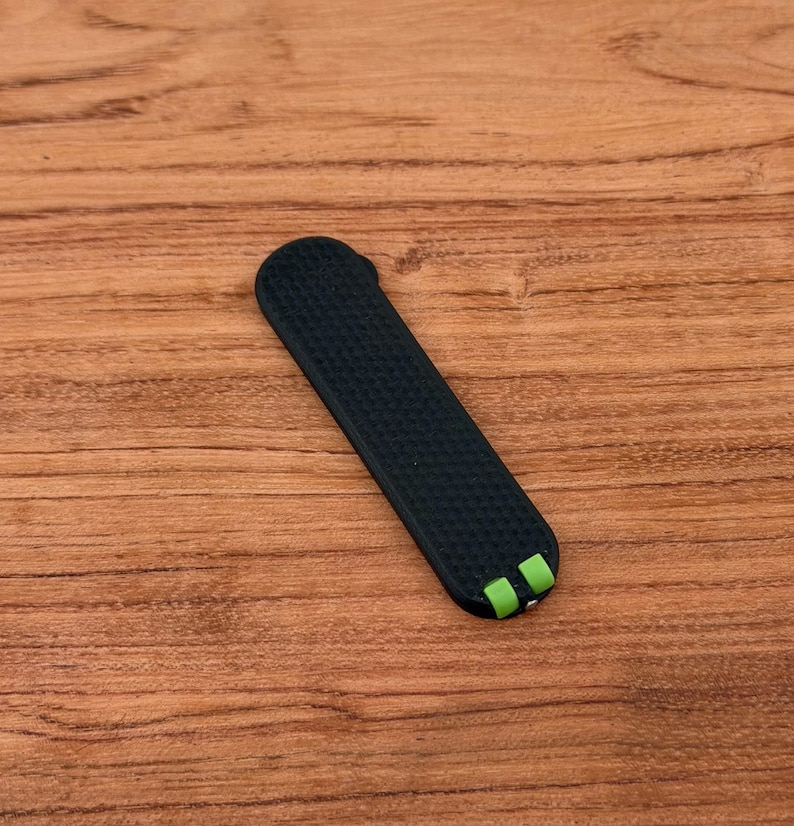 May include: A black watch band with a textured surface and green accents on the ends. The band is rectangular and appears to be made of a flexible material, resting on a wooden surface.