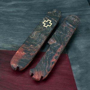 May include: Two elongated, flat wooden handle scales featuring a dark, marbled grain pattern with reddish-brown streaks. One scale displays a small, inlaid brass Swiss cross emblem near the top. Both pieces have notched ends for assembly.