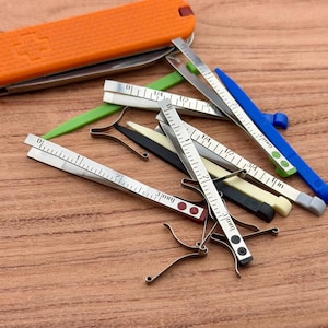 Könnte beinhalten: Ein orangefarbenes Schweizer Taschenmesser mit einem Kreuzemblem liegt neben mehreren silbernen und farbigen Messwerkzeugen. Die Werkzeuge umfassen Zoll-Lineale und verschiedene Messlehren mit grünen, blauen, schwarzen und cremefarbenen Akzenten.