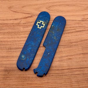 Op de afbeelding: Twee blauwe, gemarmerde mesgrepen. Eén greep heeft een gouden kruissymbool. De grepen zijn langwerpig met afgeronde uiteinden en een gebogen inkeping. De achtergrond is een houten oppervlak.