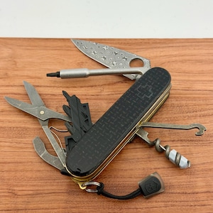 May include: A multi-tool pocket knife with a black textured handle featuring a cross emblem. The tool includes a patterned blade, a screwdriver bit, scissors, a corkscrew with a grey attachment, a hook, and a black lanyard with a small logo tag.