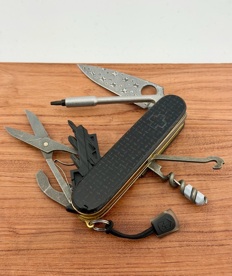 May include: A multi-tool pocket knife with a black textured handle featuring a cross emblem. The tool includes a patterned blade, a screwdriver bit, scissors, a corkscrew with a grey attachment, a hook, and a black lanyard with a small logo tag.