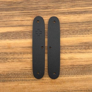 Könnte beinhalten: Zwei schwarze, strukturierte, längliche Teile mit abgerundeten Enden und jeweils drei Löchern. Ein Teil hat ein kleines Pluszeichen. Die Teile liegen auf einer Holzoberfläche.