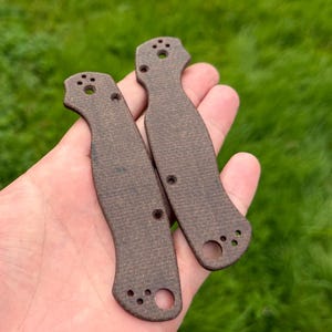 May include: Two brown knife handle scales with multiple holes for screws. The scales are made of a textured material and are designed to fit a folding knife. The background is a blurred green lawn.