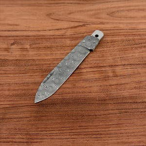 May include: A stainless steel knife blade with a decorative pattern. The blade is silver with a detailed, swirling design. The handle is also silver and has a hole at the end. The knife is set against a wooden background.