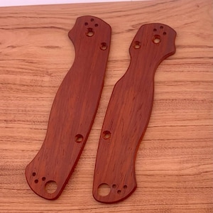 May include: Two reddish-brown wooden knife handle scales with a smooth finish. Each scale has a curved shape with multiple holes for attachment. The wood grain is visible, and the scales are displayed on a light brown wooden surface.