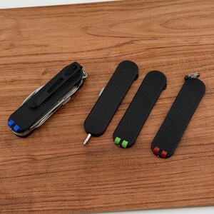 May include: A collection of black multi-tools and pocket knives with textured grips. One tool has a variety of functions, while the others are smaller, with colored accents of blue, green, and red. The tools are arranged on a wooden surface.