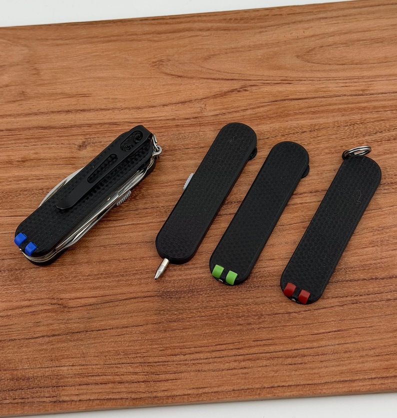 May include: A collection of black multi-tools and pocket knives with textured grips. One tool has a variety of functions, while the others are smaller, with colored accents of blue, green, and red. The tools are arranged on a wooden surface.