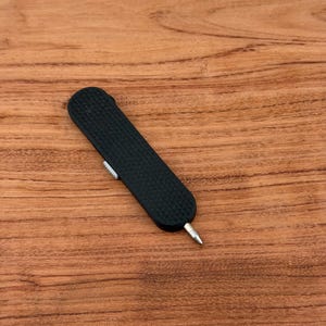 May include: A black, textured, pocket-sized tool with a silver tip and a small, white slider. The tool is rectangular with rounded ends. The background is a wooden surface.
