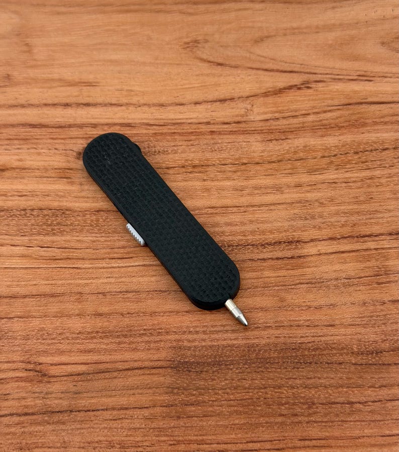 May include: A black, textured, pocket-sized tool with a silver tip and a small, white slider. The tool is rectangular with rounded ends. The background is a wooden surface.