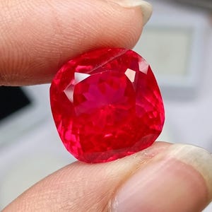 Puede incluir: Una gema vibrante de corte cojín con un color rojo profundo y saturado, con facetas intrincadas que reflejan la luz. La piedra presenta una forma cuadrada con esquinas redondeadas, mostrando una claridad brillante, transparente y pulida.