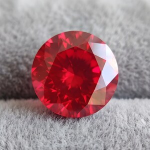 May include: A round, faceted gemstone with a vibrant, deep red color. The stone features a brilliant cut with numerous triangular and kite-shaped facets that reflect light, creating a sparkling appearance across its circular surface.