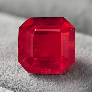 Può includere: Una gemma di colore rosso intenso e vibrante tagliata in forma ottagonale con molteplici sfaccettature che riflettono la luce. La pietra presenta una superficie chiara e lucida ed è appoggiata su una base grigia morbida e testurizzata.