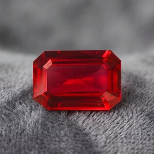 May include: A vibrant red gemstone cut into a rectangular emerald shape with faceted surfaces that reflect light. The stone features a deep, saturated crimson hue and sharp, clean edges, showcasing a clear, transparent quality throughout the piece.