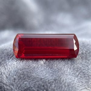 May include: A rectangular, faceted gemstone with a deep red color. The stone features a long, horizontal shape with beveled edges and a polished, translucent surface that reflects light across its multiple flat, geometric facets.