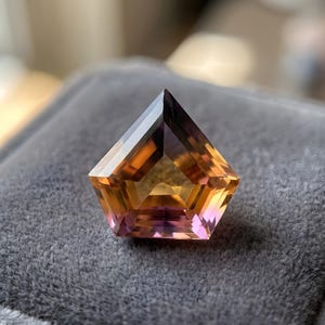 Peut inclure: Une pierre précieuse taillée en forme de pentagone. La gemme présente un dégradé de couleurs, passant du violet foncé au sommet à des nuances d'orange et de rose vers le bas. Elle repose sur un tissu de velours gris, mettant en valeur son éclat.