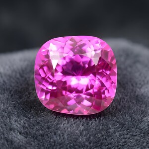 Puede incluir: Una vibrante gema rosa, de talla cojín, con múltiples facetas. La gema es de un color fucsia brillante y se encuentra sobre una superficie texturizada de color gris oscuro. La gema refleja la luz, creando un efecto brillante.