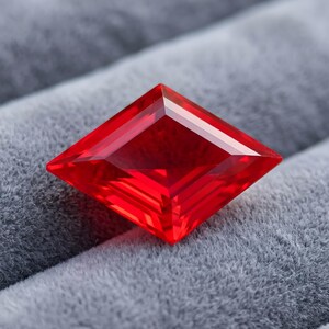 May include: A vibrant, transparent red gemstone cut into a sharp, symmetrical kite or diamond shape. The stone features multiple faceted surfaces that reflect light, creating deep crimson tones and clear geometric lines across its polished face.