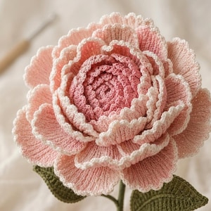May include: A handcrafted crochet flower featuring layered petals in shades of light pink and deep rose. The flower has a scalloped edge on each petal and is attached to a green stem with two dark green leaves, showcasing intricate yarn texture.