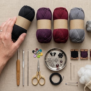 Puede incluir: Cuatro ovillos de lana en negro, morado, burdeos y gris con etiquetas de papel beige, un peque&ntilde;o carrete de hilo gris, tres ganchillos, marcadores de puntos coloridos, tijeras de metal, una bandeja con cuentas, agujas y relleno blanco.