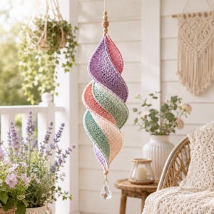 May include: A hanging spiral ornament crafted from crocheted yarn in pastel shades of purple, pink, green, and cream. The structure features a twisted design suspended by a string with wooden beads at the top and a clear faceted crystal drop at the base.