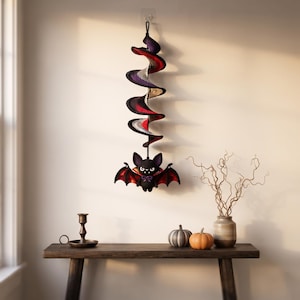 May include: A crocheted hanging decoration featuring a spiral wind spinner in black, purple, grey, and red, suspended from a hook. Attached to the bottom is a small, stuffed black bat with red-lined wings, white fangs, and a purple bow tie.