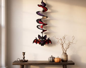 Puede incluir: Una decoración colgante de ganchillo con un molinillo de viento en espiral en negro, morado, gris y rojo, suspendido de un gancho. En la parte inferior hay un pequeño murciélago negro relleno con alas forradas en rojo, colmillos blancos y una pajarita morada.