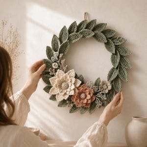 Könnte beinhalten: Ein runder Kranz aus gehäkelten salbeigrünen Blättern, der unten mit einer großen cremefarbenen Blüte und einer kleineren terrakottafarbenen Blüte verziert ist. Das Dekorationsobjekt ist aus detaillierten Garnstrukturen gefertigt und hat eine Schlaufe.