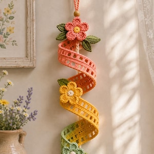 May include: A vertical wall hanging featuring a spiraling crochet ribbon in a gradient of peach, yellow, and light green. The ribbon is adorned with three crocheted flowers in matching colors, each with green leaves, and a wooden bead at the top loop.