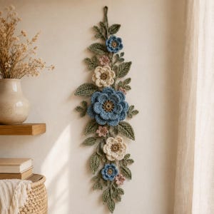 Puede incluir: Un adorno de pared vertical con flores y hojas de ganchillo en tonos azul polvoriento, crema y rosa suave. El arreglo incluye una gran flor azul central, flores más pequeñas azules y crema, y follaje verde con textura.