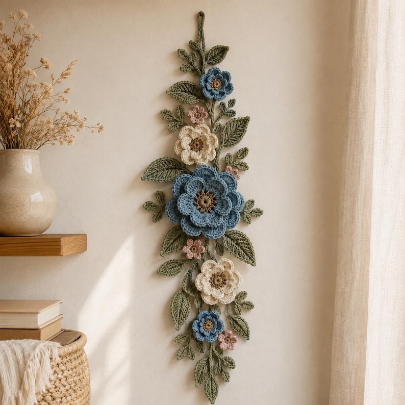Peut inclure: Une d&eacute;coration murale verticale compos&eacute;e de fleurs et de feuilles crochet&eacute;es dans des tons bleu cendr&eacute;, cr&egrave;me et rose p&acirc;le. L'arrangement comprend une grande fleur bleue centrale, des fleurs plus petites et un feuillage vert textur&eacute;.