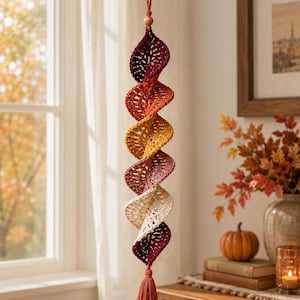 May include: A hanging crochet spiral decoration featuring a gradient of autumn-inspired colors including deep burgundy, burnt orange, golden yellow, muted rose, and cream. The piece is suspended by a string with a small wooden bead at the top.