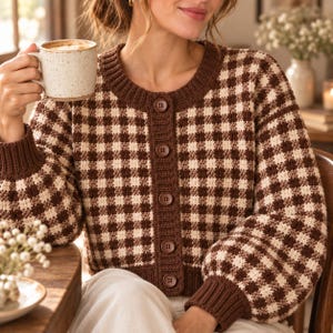 May include: A long-sleeved, button-down cardigan featuring a brown and cream checkered pattern. The garment has a solid brown ribbed crew neckline, cuffs, and a vertical button placket with five brown buttons. The knit texture is visible throughout.