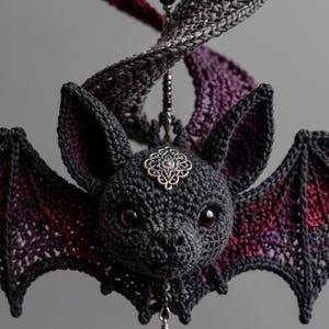 May include: A crocheted bat ornament featuring dark gray yarn with deep red and purple accents on the wings. The bat has black eyes, a silver filigree charm on its forehead, and a vertical hanging element with black beads and a red teardrop crystal.
