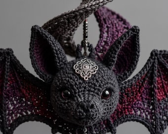Puede incluir: Un adorno de murciélago tejido a ganchillo en hilo gris oscuro con acentos en rojo profundo y morado en las alas. El murciélago tiene ojos negros, un dije de filigrana plateada en la frente y un elemento colgante con cuentas negras y un cristal.