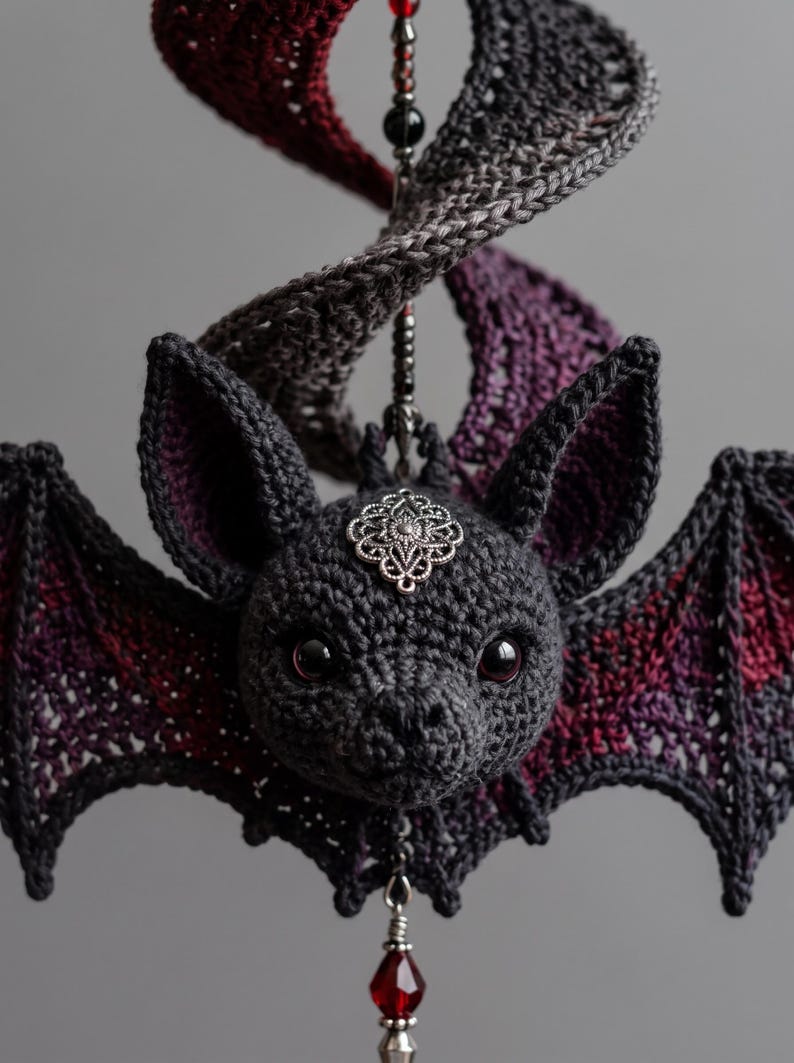 Puede incluir: Un adorno de murci&eacute;lago tejido a ganchillo en hilo gris oscuro con acentos en rojo profundo y morado en las alas. El murci&eacute;lago tiene ojos negros, un dije de filigrana plateada en la frente y un elemento colgante con cuentas negras y un cristal.