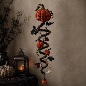 May include: A hanging crochet decoration featuring a spiral vine structure adorned with multiple orange pumpkins, dark green leaves, and a small cream-colored crescent moon charm. The piece is suspended by a black string and includes curly tendrils.