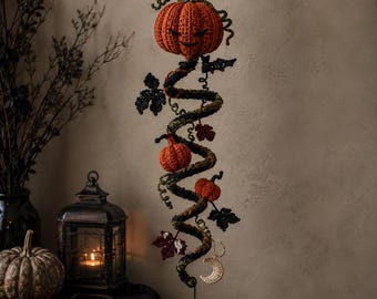 Puede incluir: Una decoración colgante de ganchillo con una estructura de enredadera en espiral adornada con varias calabazas naranjas, hojas de color verde oscuro y un pequeño dije de media luna de color crema. La pieza cuelga de un hilo negro.