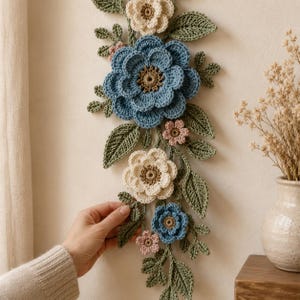 Peut inclure: Une d&eacute;coration murale verticale compos&eacute;e de fleurs au crochet dans des tons bleu, cr&egrave;me et rose p&acirc;le, agr&eacute;ment&eacute;e de feuilles et de tiges crochet&eacute;es vertes. L'ensemble comprend plusieurs fleurs superpos&eacute;es avec des c&oelig;urs textur&eacute;s.