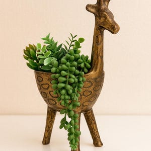 May include: A bronze-colored giraffe-shaped planter filled with green succulents. The planter has a textured surface and is supported by three legs. The giraffe's head and neck extend upwards, creating a unique decorative piece.