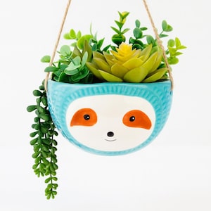 May include: A hanging planter with a sloth face design. The turquoise ceramic pot holds a variety of green and yellow faux succulents. The sloth's face has orange accents and a smiling mouth. The planter hangs from a natural rope.