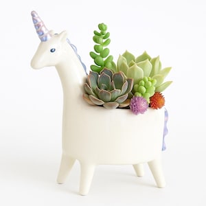 May include: A white ceramic unicorn planter with a blue and purple horn and mane. The planter is filled with a variety of succulents and small, colorful flowers. The unicorn has a whimsical design and is standing on four legs.