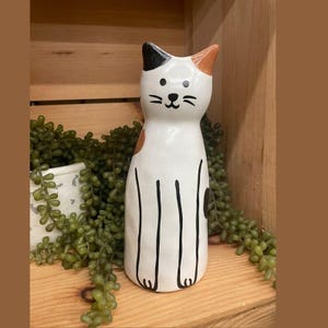 May include: A white ceramic cat figurine with black and brown markings. The cat has a smiling face, black whiskers, and black lines for legs. The figurine is a decorative item.