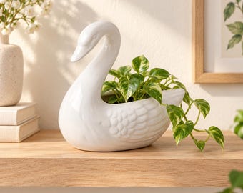 May include: A white ceramic swan-shaped planter featuring a smooth, glossy finish and molded feather details on the body. The hollow center of the swan holds a small green and yellow variegated pothos plant with trailing vines extending outward.