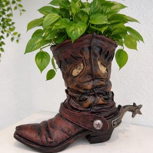 May include: A decorative planter shaped like a weathered brown cowboy boot, filled with vibrant green leafy plants. The boot features intricate details, a spur, and a strap with a silver buckle, creating a rustic, Western-themed aesthetic.