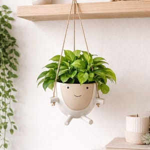 May include: A white ceramic hanging planter with a smiling face design, filled with vibrant green leafy plants. The planter is suspended by a natural rope and adds a touch of whimsy to any indoor space. The planter has a beige face and white body.