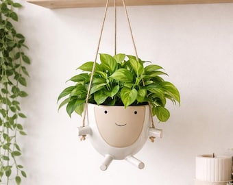 May include: A white ceramic hanging planter with a smiling face design, filled with vibrant green leafy plants. The planter is suspended by a natural rope and adds a touch of whimsy to any indoor space. The planter has a beige face and white body.