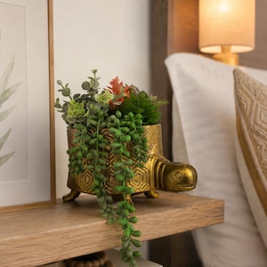 May include: A gold-colored turtle-shaped planter with a textured geometric shell pattern, containing a variety of artificial green and reddish succulents, including trailing string-of-pearls foliage that hangs over the front edge of the turtle.