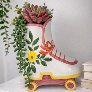 May include: A white ceramic roller skate planter with pink and yellow accents, filled with a red succulent and trailing green plants. The planter features painted floral details and a decorative bow.
