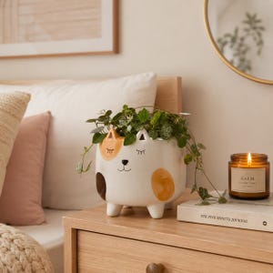 May include: A white ceramic cat-shaped planter with painted orange and brown spots, closed eyes, and a small nose. The planter features four short legs and contains a trailing green plant with small heart-shaped leaves, resting on a wooden surface.